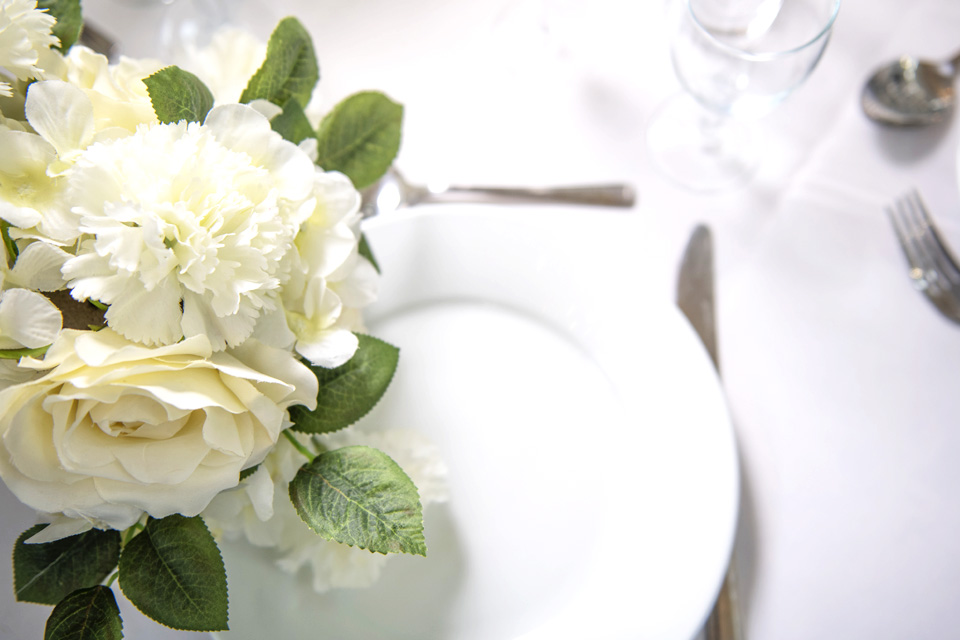 Flowers rest on a table next to a plate and cutlery.